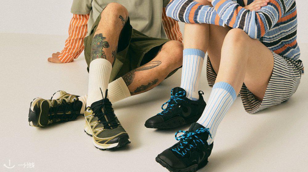 Deux personnes assises par terre, portant des pulls colorés, des shorts, des chaussettes rayées et des baskets, l'une d'elles ayant des tatouages sur les jambes.