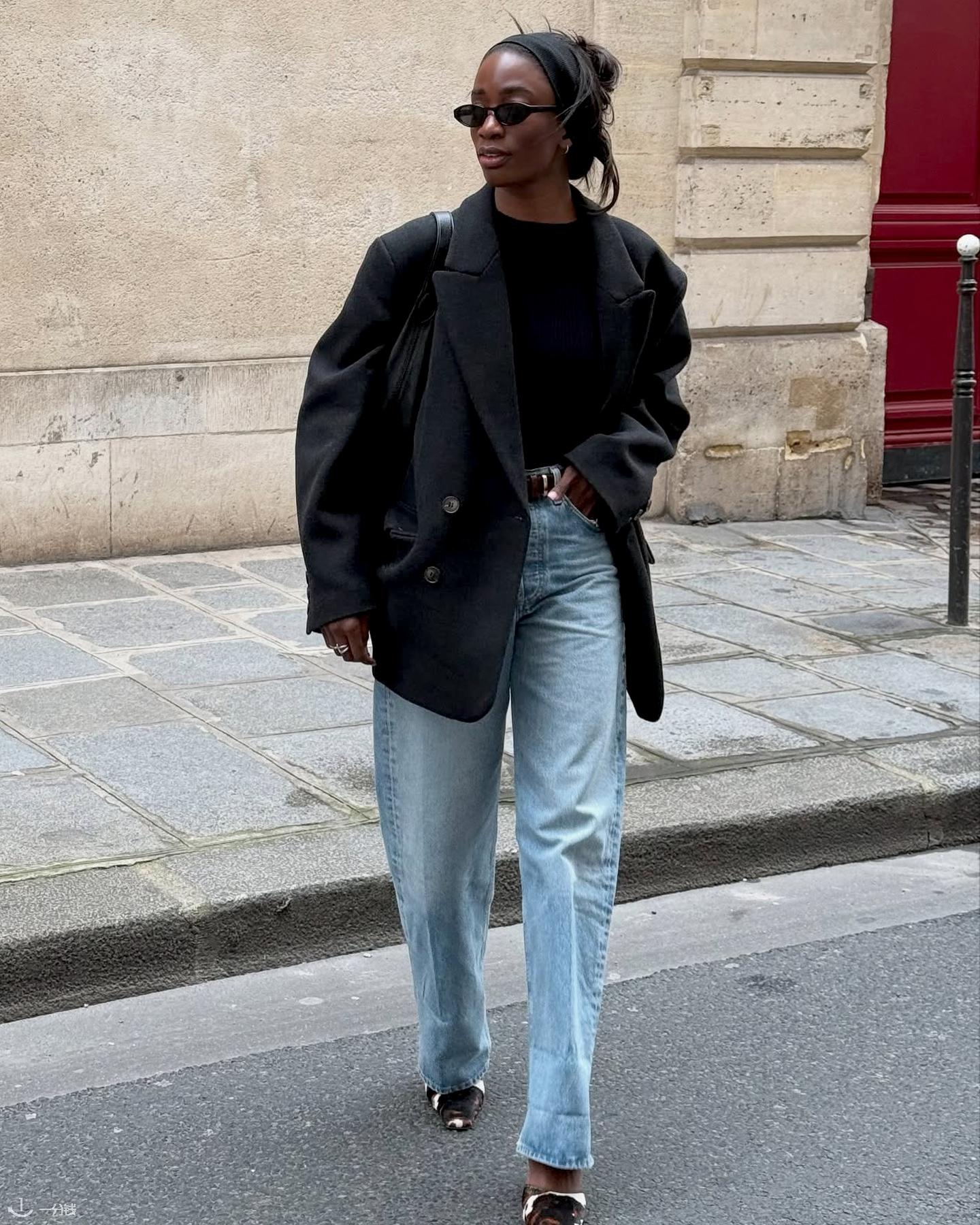 British style influencer Liv Madeline crosses a street in Paris wearing a black headband, black oval sunglasses, an oversize black blazer jacket, black top, black belt, light-wash barrel jeans, and cow-print mule heels